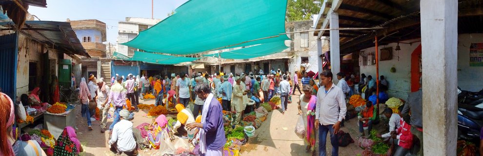 Crowded outdoor market street in Varanasi with people in traditional Indian clothing, vendors selling orange and yellow marigold flowers under green tarpaulin shade covers