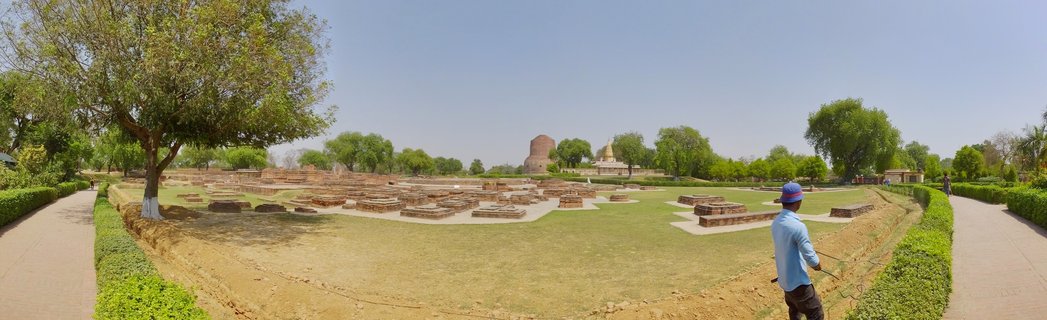 Panoramic view of ancient Buddhist archaeological site showing brick ruins, Dhamek Stupa tower, green lawns, trees, and a worker in blue shirt