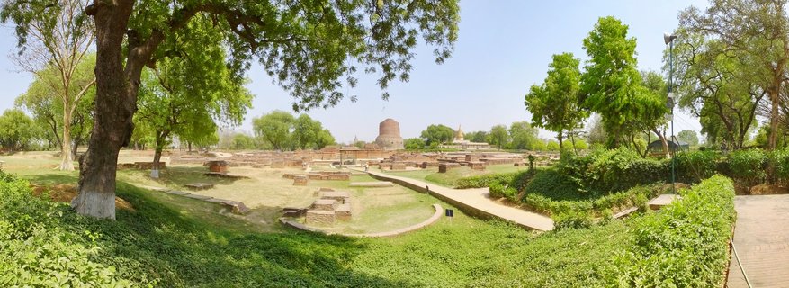Panoramic view of the Dhamek Stupa and archaeological ruins at Sarnath Buddhist site with green lawns, trees, and walking paths under clear blue sky