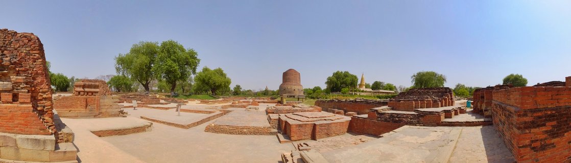 Panoramic view of ancient red brick Buddhist ruins with the cylindrical Dhamek Stupa at center, surrounded by archaeological foundations, green trees, and clear blue sky at Sarnath, India.