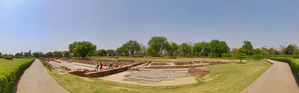 Panoramic view of ancient brick archaeological ruins at Sarnath with visitors walking among the foundations, surrounded by green lawns and trees under clear blue sky