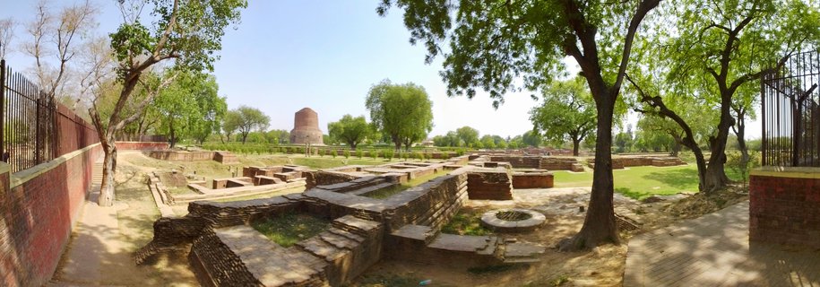 Panoramic view of ancient brick monastery ruins with the Dhamek Stupa visible in the distance at Sarnath Buddhist pilgrimage site, surrounded by green trees under clear blue sky
