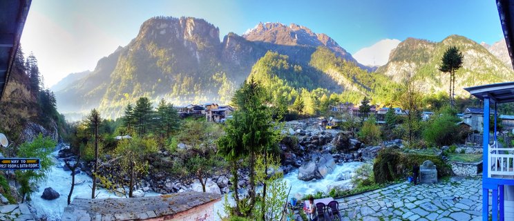 Panoramic view of a mountain village in Nepal with a rushing river, pine trees, traditional buildings on hillsides, and snow-capped Himalayan peaks in the background under clear blue sky