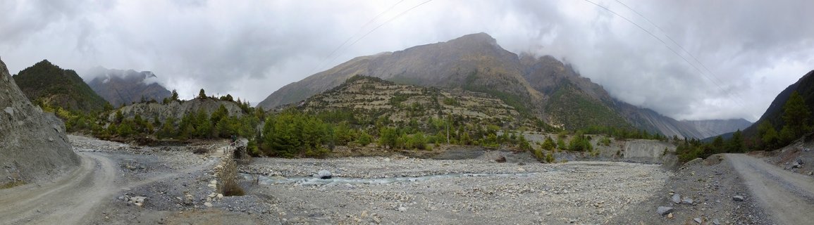 Panoramic mountain landscape in Nepal showing a rocky riverbed in the foreground with dirt trekking paths on either side. Terraced hillsides with sparse pine trees and shrubs rise toward a large mountain peak partially covered by clouds. Overcast sky above the Annapurna Circuit region.