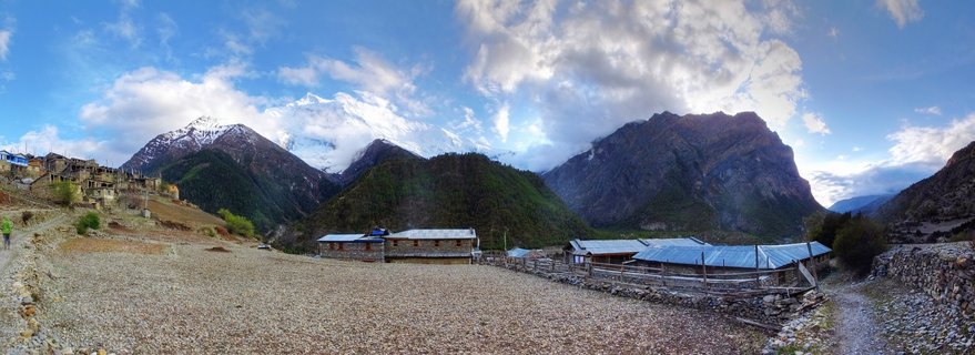 Wide mountain landscape showing stone buildings in a gravel valley with snow-covered Himalayan peaks and blue sky with clouds in Nepal