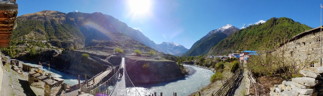Wide panoramic view of a suspension bridge crossing a turquoise river in a Himalayan valley with snow-capped mountains, stone buildings, and green terraced hillsides in Pisang, Nepal