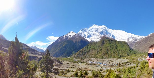 Wide mountain landscape showing a valley village with terraced fields below snow-covered Himalayan peaks under clear blue sky