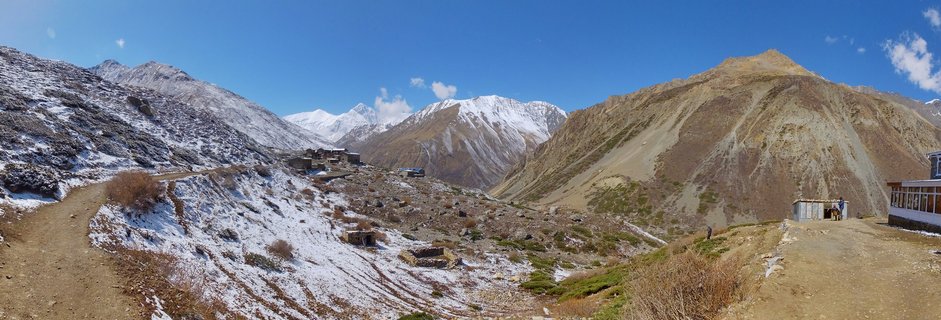 Panoramic mountain landscape showing Yak Kharka settlement with stone buildings on snow-patched terrain, surrounded by steep Himalayan slopes and distant snow-capped peaks under blue sky