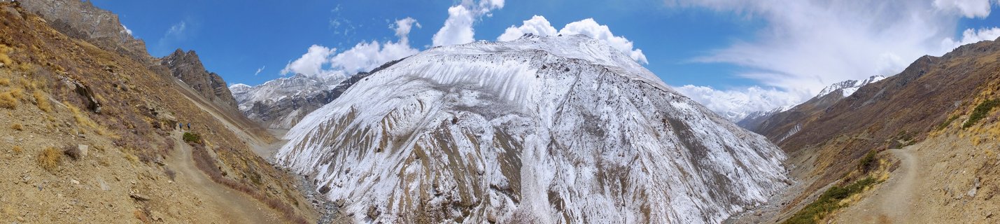 Panoramic view of a snow-covered mountain face in Yak Kharka valley with dirt trekking trails on both sides, blue sky with white clouds, Annapurna region, Nepal