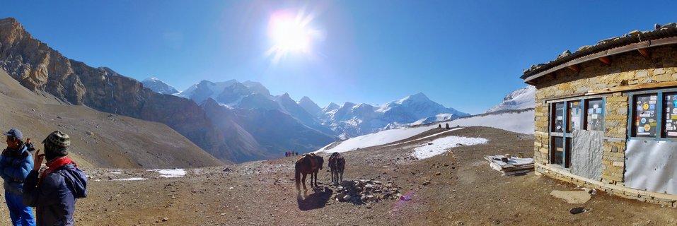 Mountain landscape showing trekkers, horses, and a stone building with snow-capped Himalayan peaks in the background under a bright sun and clear blue sky