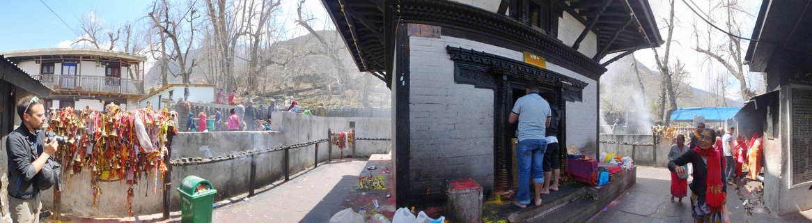 Wide view of Muktinath Temple showing white-walled shrine with dark wood trim, pilgrims in colorful clothing entering the sacred building, prayer flags hanging on stone walls, mountains visible in background under clear blue sky