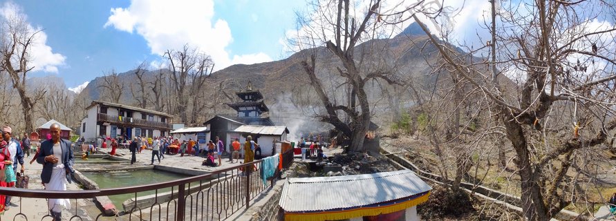 Wide panoramic view of Muktinath Temple complex in Nepal showing pilgrims around sacred water pools, traditional pagoda-style temple buildings, bare trees, and mountain landscape under blue sky
