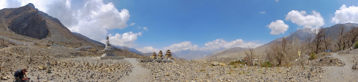 Panoramic landscape showing a large white Buddhist stupa with golden spires in the Muktinath region of Mustang, Nepal. Three smaller golden-topped stupas surround the main structure. Rocky high-altitude terrain with sparse vegetation and leafless trees. Himalayan mountain range visible in the background under blue sky with white clouds. Prayer flags and one person visible in the scene.