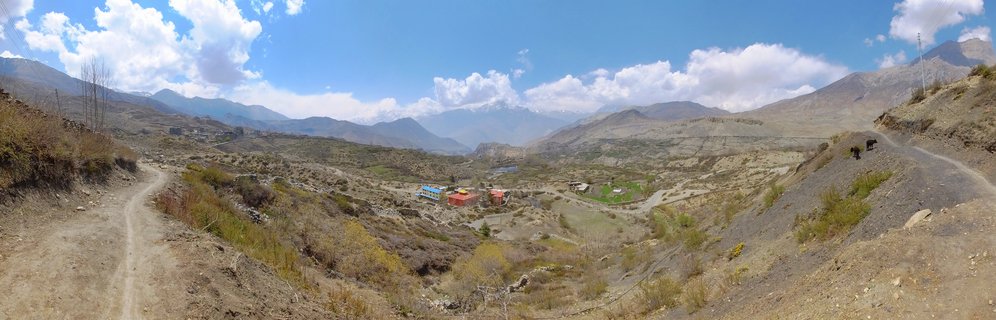 Wide panoramic view of Muktinath-Kagbeni valley in Nepal showing traditional buildings with colorful roofs in an arid high-altitude desert landscape, winding dirt roads, two pack animals on a hillside path, distant snow-capped Himalayan mountains, and blue sky with white clouds