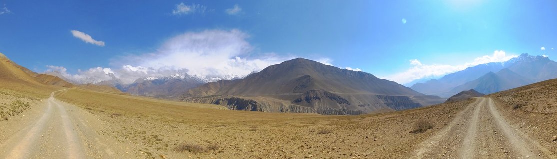 Panoramic landscape showing two dirt roads diverging across arid brown terrain with barren mountains and snow-capped Himalayan peaks in the distance under blue sky with scattered clouds