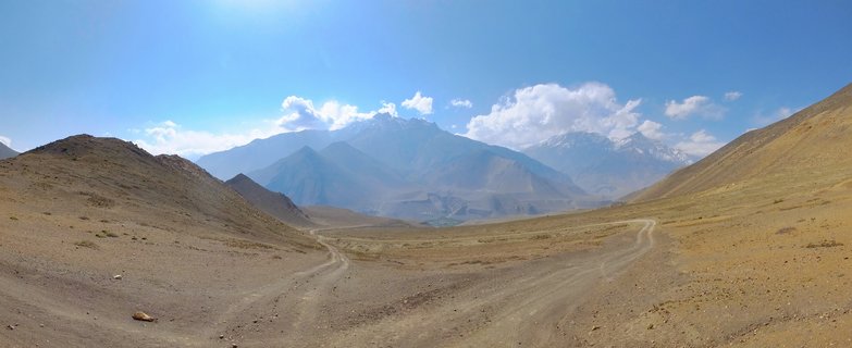 Wide mountain valley with winding dirt road through arid brown terrain leading to distant snow-capped Himalayan peaks under blue sky with white clouds
