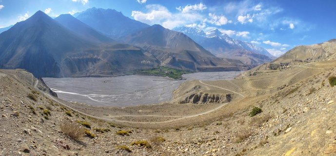 Panoramic mountain landscape showing a wide valley with a braided river, surrounded by barren brown mountains with snow-capped Himalayan peaks in the distance under a blue sky with scattered clouds
