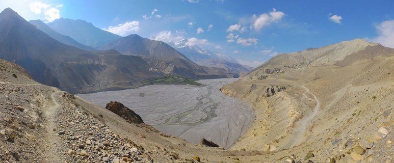 Wide valley landscape showing a braided river flowing through arid mountains in Mustang, Nepal, with dirt roads winding along the hillsides and snow-capped peaks visible in the distance under a blue sky