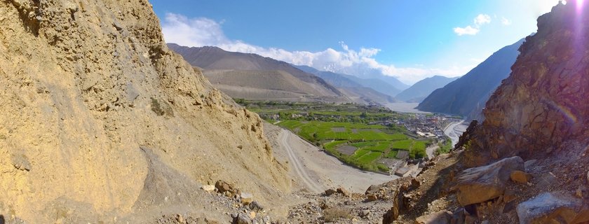 Mountain valley landscape showing rocky cliffs framing a green agricultural valley with a river, village settlement, and snow-capped Himalayan peaks under blue sky