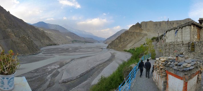 Wide valley landscape showing a braided riverbed between barren mountains in Mustang, Nepal, with two people walking on a path beside a stone building with prayer flags