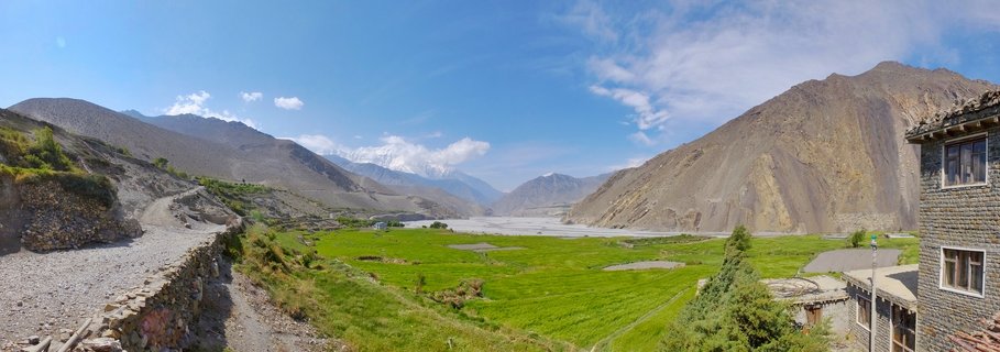 Wide panoramic landscape showing green valley floor with stone buildings, surrounded by barren brown mountains and distant snow-capped peaks under blue sky in Mustang, Nepal
