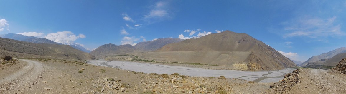 Wide panoramic view of Kali Gandaki river valley in Nepal showing a braided riverbed between barren brown mountains with snow-capped Himalayan peaks in the distance under blue sky with white clouds