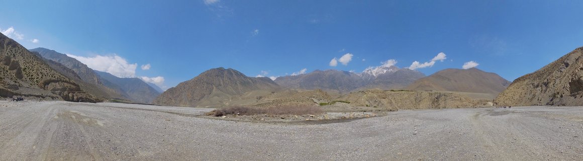 Wide mountain valley landscape in Nepal showing rocky riverbed, brown mountain slopes, and snow-capped Himalayan peaks under blue sky with white clouds