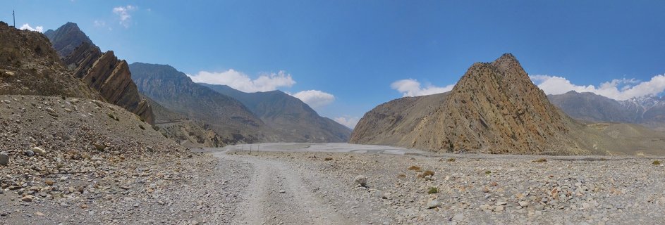 Wide mountain valley in Nepal with rocky terrain, a dirt road leading through the center, steep brown mountains on both sides, and snow-capped peaks visible in the distance under a blue sky with scattered clouds