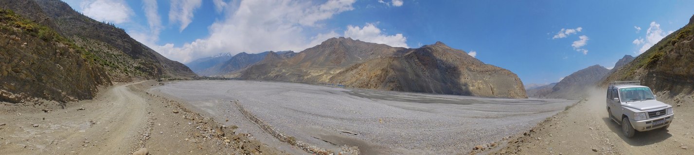 Panoramic view of a white SUV driving on a dirt road through a wide rocky riverbed valley with barren mountains on both sides under blue sky with clouds in Mustang, Nepal