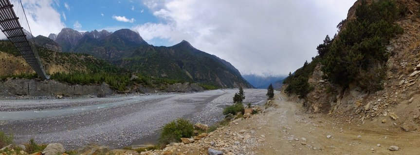 Wide river valley in Nepal with braided Kali Gandaki River, partial suspension bridge on left, dirt path along hillside, forested mountains and snow-capped Himalayan peaks in background under cloudy sky