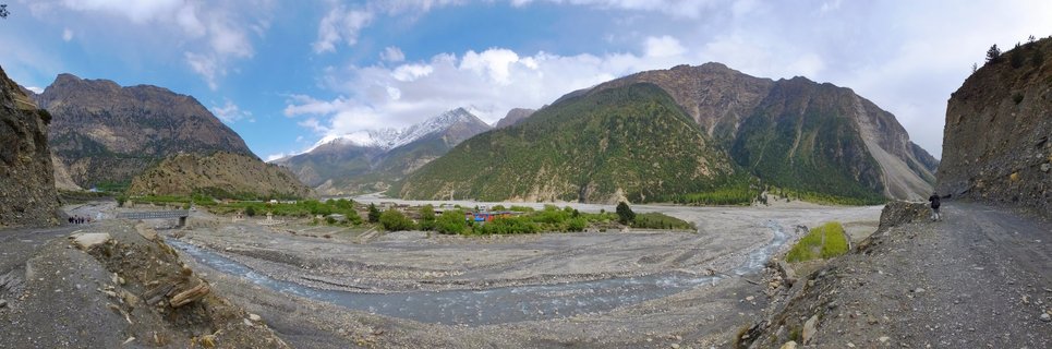Wide mountain valley in Nepal with a winding river, green slopes, rocky mountains, and snow-capped peaks in the distance under blue sky with clouds