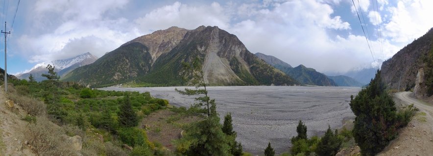 Panoramic mountain landscape showing the Kali Gandaki river valley in Nepal with a prominent rocky mountain peak, gray braided riverbed with stones, green coniferous trees in the foreground, and overcast cloudy sky