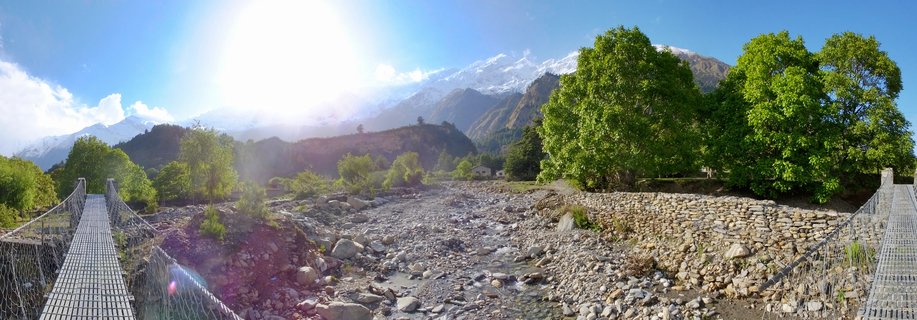 Steel suspension bridge crossing a rocky riverbed with snow-capped Himalayan mountains and green trees under bright blue sky in Nepal