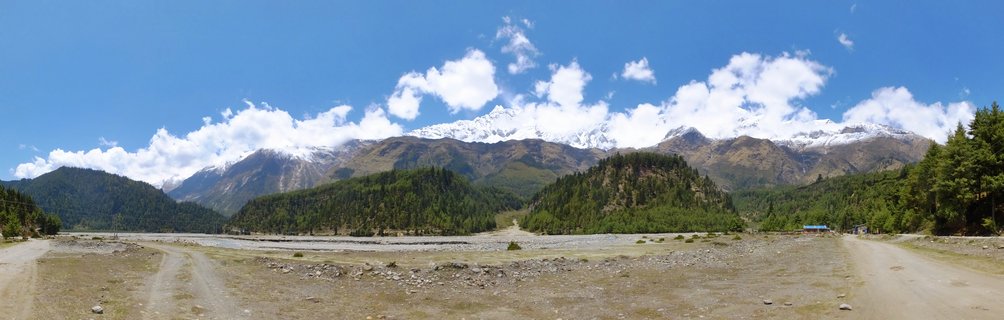 Wide mountain landscape showing snow-capped Himalayan peaks above green forested hills and a rocky valley floor with dirt roads under blue sky with white clouds