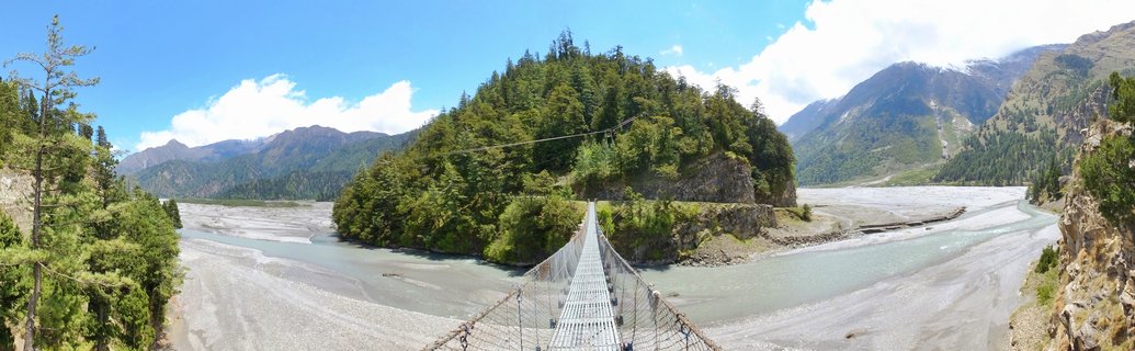 Panoramic view of a metal suspension bridge extending toward a forested island in a wide braided river valley, with snow-capped Himalayan mountains and blue sky with clouds, Larjung-Ghasa, Nepal