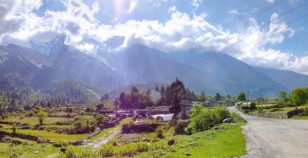 Panoramic landscape showing snow-capped Himalayan mountains above green terraced fields, stone walls, village buildings, and a winding dirt road in Larjung, Nepal