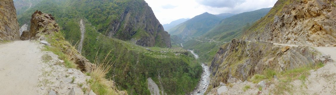 Wide mountain valley view showing a dirt road carved into rocky cliff sides with a river flowing through green slopes below under cloudy sky