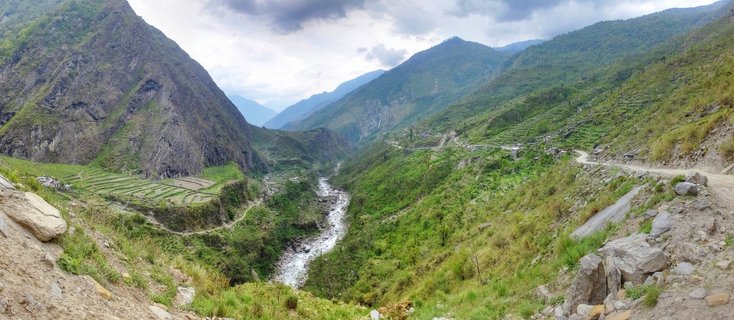 Panoramic view of a deep mountain valley in Nepal with a river running through it, green terraced fields on hillsides, steep rocky mountains, and a winding dirt road on the right slope under cloudy sky
