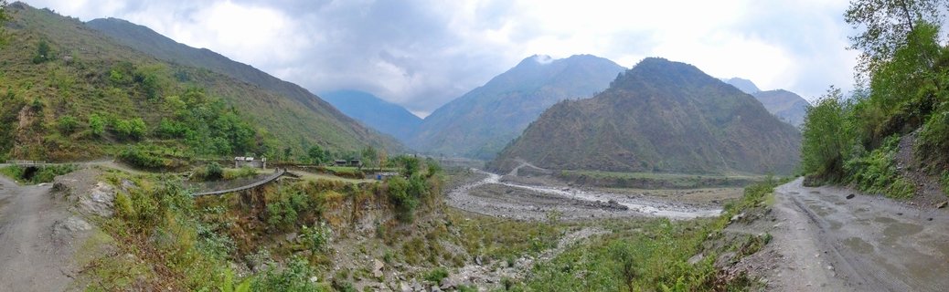 Wide mountain valley in Nepal showing a riverbed running through green slopes with dirt roads on both sides and cloudy sky above the peaks