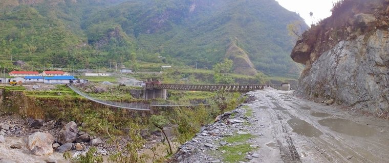 Mountain valley landscape showing a suspension bridge crossing a rocky river, with buildings featuring blue and red roofs on the left bank, a muddy road along a cliffside on the right, and steep green mountains in the background
