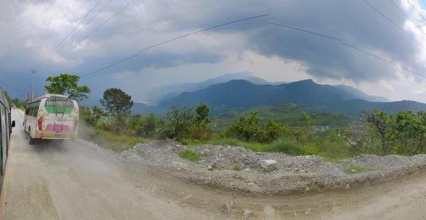 A colorful bus driving on a dusty mountain road with green hills and mountain ranges in the background under cloudy skies in Nepal