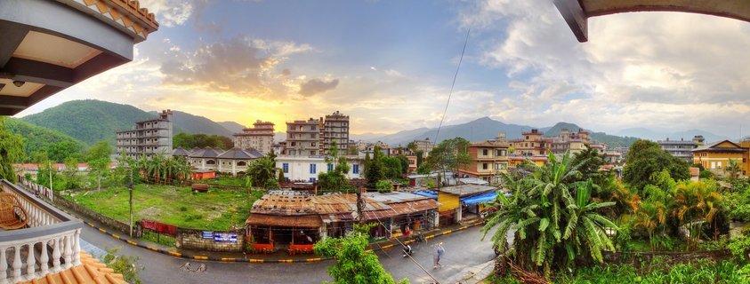 Panoramic cityscape of Pokhara, Nepal at sunset showing buildings, tropical trees, streets, and mountains in the background under a cloudy golden sky