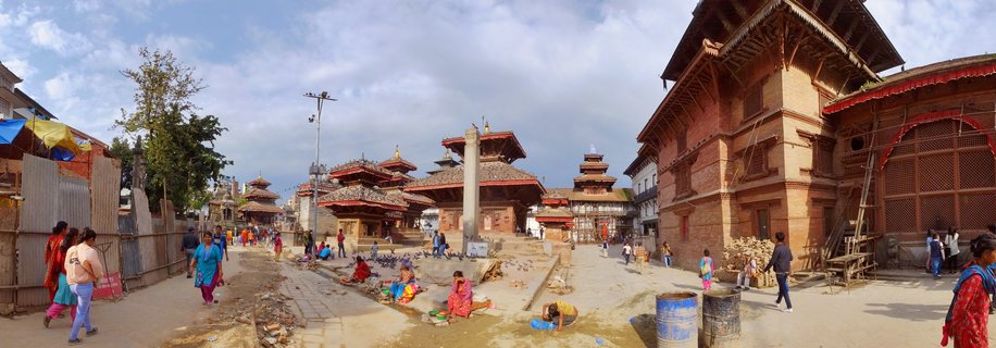 Wide panoramic view of traditional Nepalese pagoda temples in Kathmandu Durbar Square with red brick buildings, people in colorful clothing walking and sitting in the plaza, blue sky with scattered clouds above