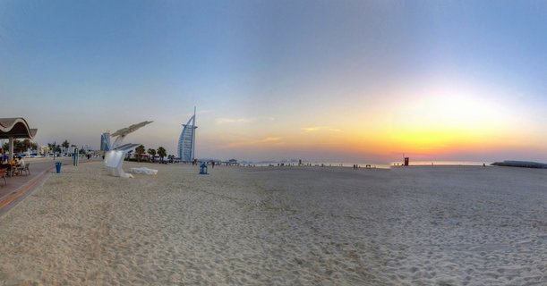 Panoramic view of Burj Al Arab hotel at sunset from Jumeirah Beach, with white sand foreground, beach visitors, modern sculptures, and golden-orange sky over the Arabian Gulf
