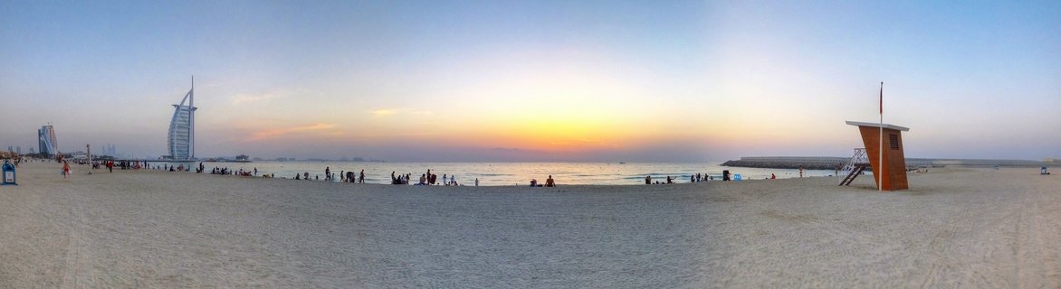 Panoramic view of Burj Al Arab hotel at sunset from a sandy beach with people scattered along the shoreline and a lifeguard tower on the right