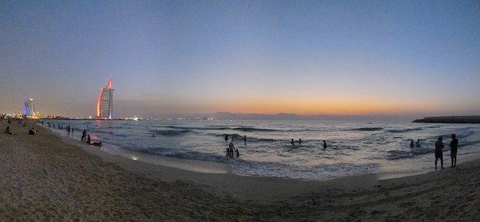 Panoramic beach scene at dusk showing the illuminated Burj Al Arab hotel in the distance with people swimming in the ocean and walking on the sand