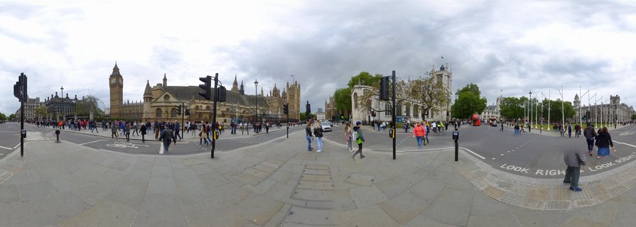 Wide panoramic view of Parliament Square showing Elizabeth Tower (Big Ben), Palace of Westminster, Westminster Abbey towers, pedestrians on stone pavement, traffic lights, black poles, and 'LOOK RIGHT' road markings under overcast sky.