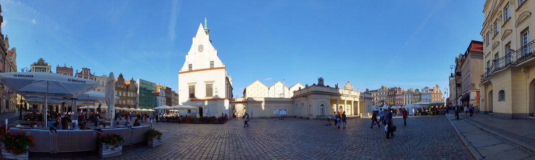 Wide view of Poznan Old Market Square showing white Town Hall building with clock tower, cobblestone plaza, outdoor cafe seating with umbrellas, and colorful historic buildings under blue sky