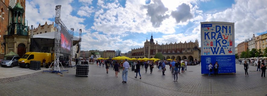 Wide panoramic view of Krakow Main Market Square showing St Mary's Basilica brick Gothic church, Renaissance Cloth Hall, yellow vendor umbrellas, and crowds of tourists on cobblestone plaza under blue sky with clouds