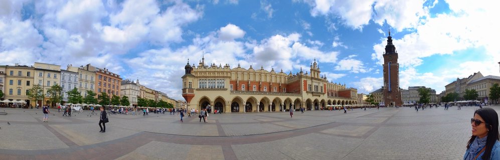 Wide view of Krakow's Main Market Square showing the Cloth Hall building in center, St. Mary's Basilica tower on right, historic colorful buildings, cobblestone plaza with tourists, blue sky with white clouds